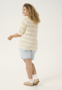 Pull en crochet beige à manches courtes, présentant un motif rayé. Associé à un short en jean bleu clair et des baskets beiges.