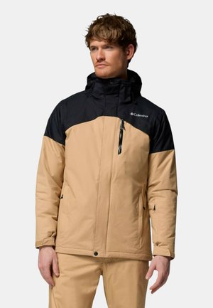 Uomo che indossa una giacca Columbia beige e nera con cappuccio, in piedi e che guarda leggermente di lato su uno sfondo semplice.