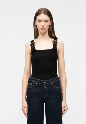 Νεαρή γυναίκα με ίσια καστανά μαλλιά, που φοράει μαύρο τοπ χωρίς μανίκια με διακοσμημένους ιμάντες και σκούρα μπλε τζιν ψηλής μέσης, κοιτάζοντας εμπρός.