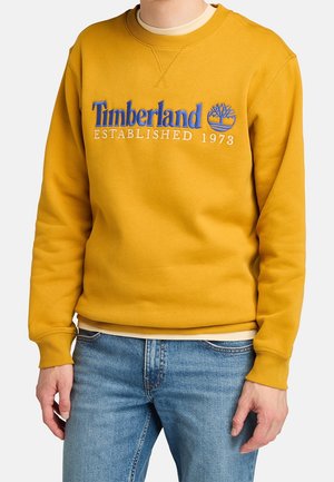 Mężczyzna w musztardowożółtej bluzie Timberland z niebieskim logo i jasnoniebieskich dżinsach, stojący na jednolitym tle.