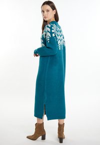 Langes türkisfarbenes Strickkleid mit weißem Blattmuster, hohem Halsausschnitt und Schlitz am Saum. Aus weichem, strukturiertem Material gefertigt.
