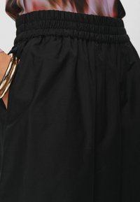 Pantalon noir avec taille élastique, comportant des poches latérales et un tissu lisse légèrement texturé. Accents en métal doré visibles.
