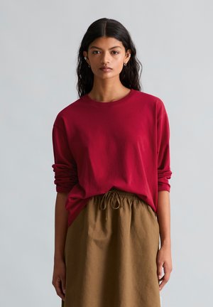 Jeune femme aux longs cheveux foncés porte une chemise rouge ample à manches longues et une jupe marron à cordon sur un fond gris uni.