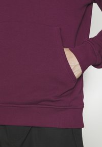 Main dans la poche avant d'un sweat violet à manches longues avec poignets et ourlet côtelés, porté avec un pantalon noir.