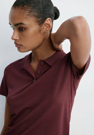 Jeune femme avec les cheveux en chignon, portant un polo bordeaux, regardant vers le bas avec une main touchant l'arrière de son cou.