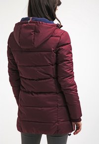 Bordowa puchowa kurtka z kapturem, z poziomymi pikowanymi sekcjami, elastycznymi mankietami i niebieskim akcentem przy kołnierzu.