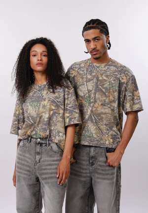 Deux jeunes adultes se tiennent côte à côte, portant des t-shirts camouflage assortis et des jeans gris, sur un fond clair uni.