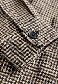 Tessuto a motivo pied-de-poule in marrone, crema e nero; presenta un bottone nero. La texture sembra morbida e tessuta, con un colletto strutturato.