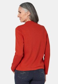 Rode, geribbelde gebreide cardigan met lange mouwen en een kraag. Gecombineerd met donkere jeans met decoratieve stiksels op de achterzakken.