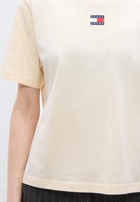 Μπεζ βαμβακερό t-shirt με κοντά μανίκια, που διαθέτει ένα μικρό μπλ ε σκούρο και κόκκινο αυτοκόλλητο με λογότυπο στο στήθος. Μαλακή υφή και άνετη εφαρμογή.