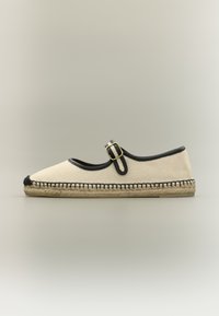 Espadrille en toile beige avec bordure noire, bride à boucle sur le dessus, pointe cousue noire et semelle en jute tissée sur fond neutre.