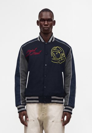 Mann trägt eine marineblaue Varsity-Jacke mit grauen Ärmeln, gestreiftem Kragen und Bündchen, besticktem Astronautengesicht auf der rechten Brust und rotem Text auf der linken Brust.