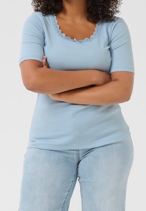 Kvinde med krøllet hår iført en lyseblå kortærmet skjorte og lyseblå jeans, stående med armene krydsede mod en hvid baggrund.