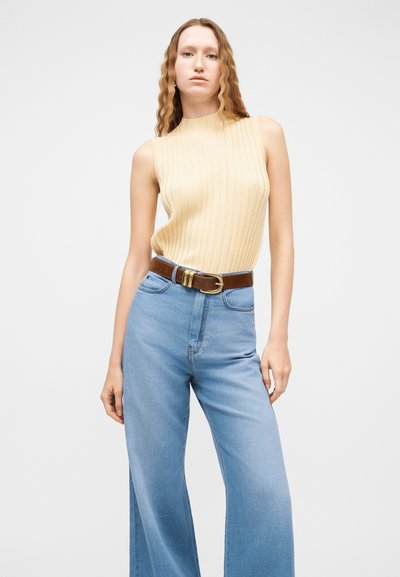 Femme aux cheveux longs et ondulés portant un haut sans manches beige à côtes, un jean bleu clair taille haute à jambes larges et une ceinture marron avec une boucle en or.