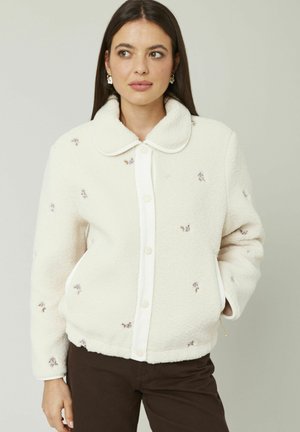 Veste crème floue avec une texture douce, un col arrondi, des boutons sur le devant et des accents de broderie florale sur l'ensemble.