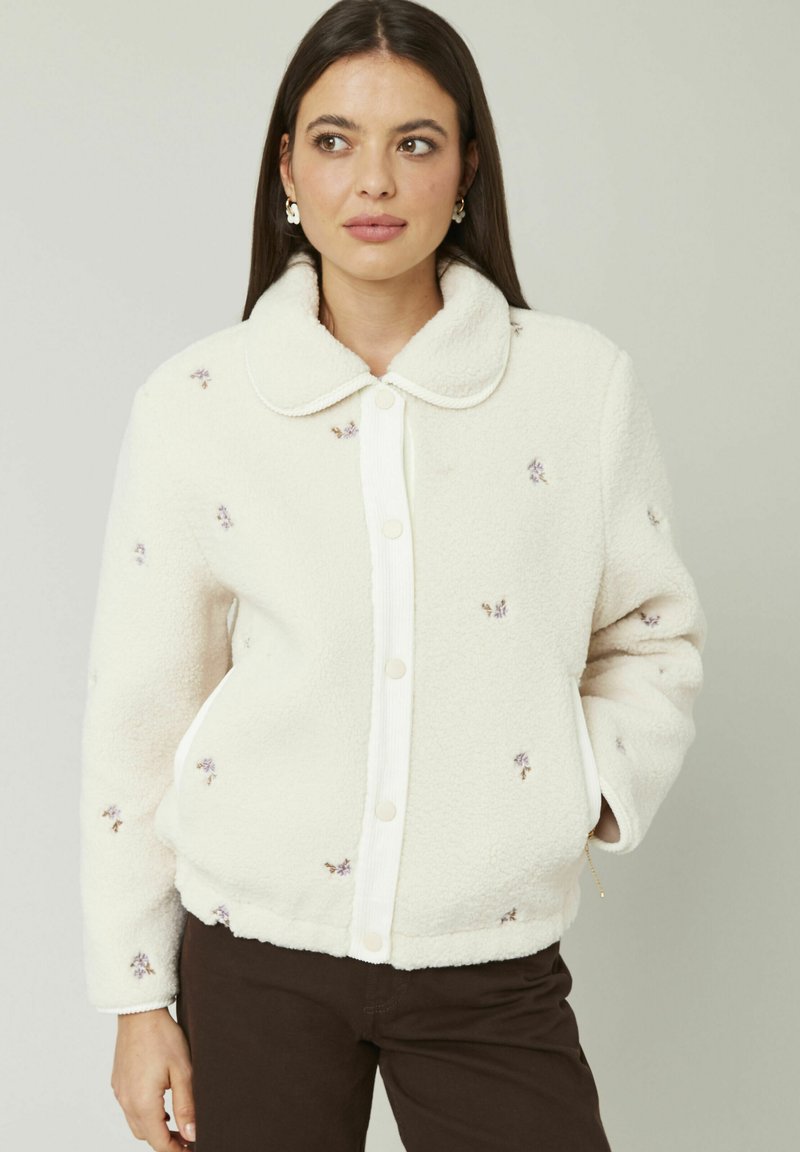 Veste crème floue avec une texture douce, un col arrondi, des boutons sur le devant et des accents de broderie florale sur l'ensemble.