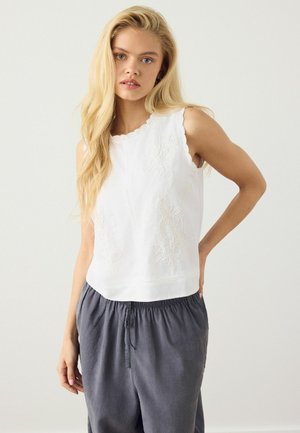 Young woman with long blonde hair wearing a sleeveless white embroidered top and loose gray pants against a plain background.