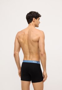 Giovane uomo con capelli corti e scuri che indossa slip neri con elastico blu HUGO, visto da dietro su uno sfondo chiaro e semplice.