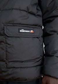 Gros plan sur une veste noire matelassée montrant une poche avec un écusson du logo ellesse et une partie de la main et du bras d'une personne.