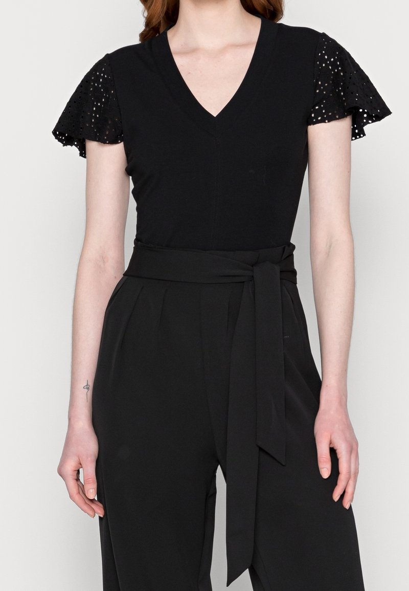 Femme portant un haut noir à col en V avec des manches volantées ajourées et un pantalon noir taille haute avec une ceinture en tissu nouée.