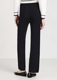 Person, der hochtaillierte schwarze Hosen, ein weißes Langarmoberteil mit schwarzem Saum und schwarze Flachschuhe trägt, steht vor einem schlichten Hintergrund.