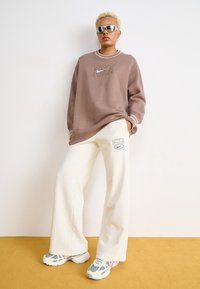 Túlméretezett barna pulóver fehér részletekkel és Nike logóval, krémszínű, bő szárú melegítőnadrággal, amelyen márkajelzés található, és színes sportcipőkkel.