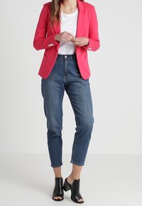 Blazer rose à revers crantés et boutons argentés, associé à une chemise blanche et un jean bleu taille haute. Mules noires à talons ouverts.