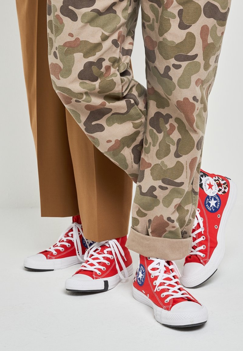 Sneaker alte in tela rossa con suole e lacci bianchi. Abbinati a pantaloni beige camouflage e pantaloni larghi marroni.