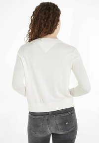 Maglione bianco lavorato a maglia con maniche lunghe, colletto e orlo a coste, caratterizzato da un dettaglio di design triangolare sul retro, abbinato a jeans grigi scuri.