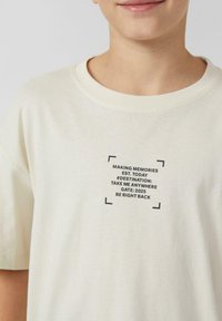 T-shirt en coton couleur crème avec un motif imprimé noir et un texte dans un design carré. Manches courtes et col rond.
