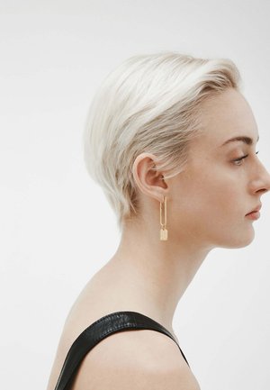 Pendientes de oro con ganchos alargados y charms rectangulares. La modelo tiene el pelo corto y rubio, y lleva una blusa negra que resalta los pendientes.