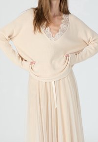 Pull beige en maille avec un large col en V et une bordure en dentelle, associé à une jupe fluide et transparente de couleur crème, ornée d'un ruban noué à la taille.
