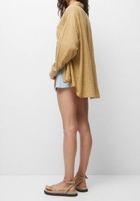 Chemise oversize jaune moutarde à manches longues, tissu texturé et coupe décontractée, associée à un short en denim et des sandales beiges à lanières.