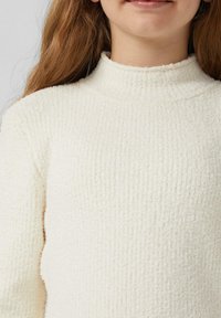 Person mit langen braunen Haaren, die einen strukturierten, cremefarbenen Strickpullover mit hohem Kragen trägt und sanft lächelt.
