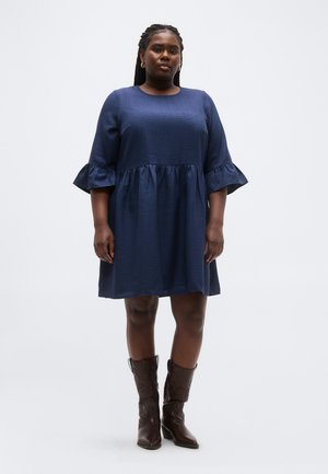 Žena s pletenými vlasmi, oblečená v tmavomodré šaty po kolená s volánikovými rukávmi a hnedými čižmami do polovice lýtok, stojaca pred jednofarebným pozadím.