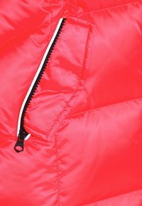 Le tissu du manteau matelassé rouge avec une texture lisse présente une poche zippée noire, avec des accents blancs contrastants le long de la fermeture éclair.