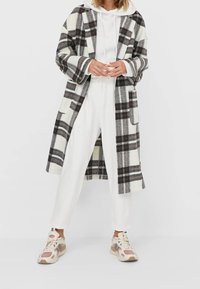 Lange plaidjas in het wit, zwart en bruin, met een capuchon en twee voorzakken, gecombineerd met witte joggers en multikleuren sneakers.