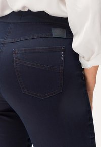 Jeans di colore blu scuro con vita media, tasca posteriore con elemento decorativo costituito da due borchie argentate e cuciture a contrasto.
