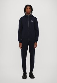 Jūras spēku zip-up kapuce ar saites un logotipu uz krūtīm, saskaņota ar atbilstošām jogger biksēm. Gluda auduma un pieguļoša dizaina; valkāta ar melniem tenisa apaviem.