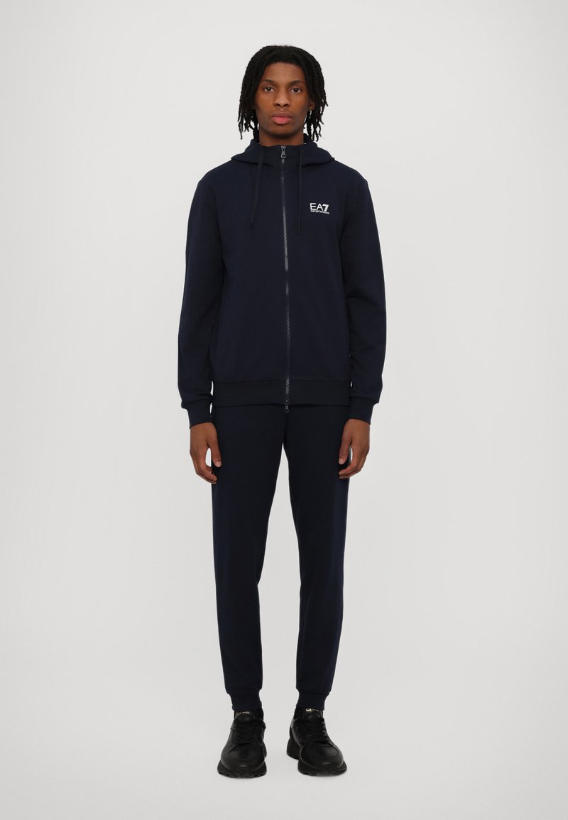 Jūras spēku zip-up kapuce ar saites un logotipu uz krūtīm, saskaņota ar atbilstošām jogger biksēm. Gluda auduma un pieguļoša dizaina; valkāta ar melniem tenisa apaviem.