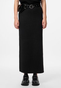 Črna rebrasta maxi krila s prilegajočim silhueto, v kombinaciji z svetlečo črno pasom in črnimi športnimi čevlji. Gladek, teksturiran material.