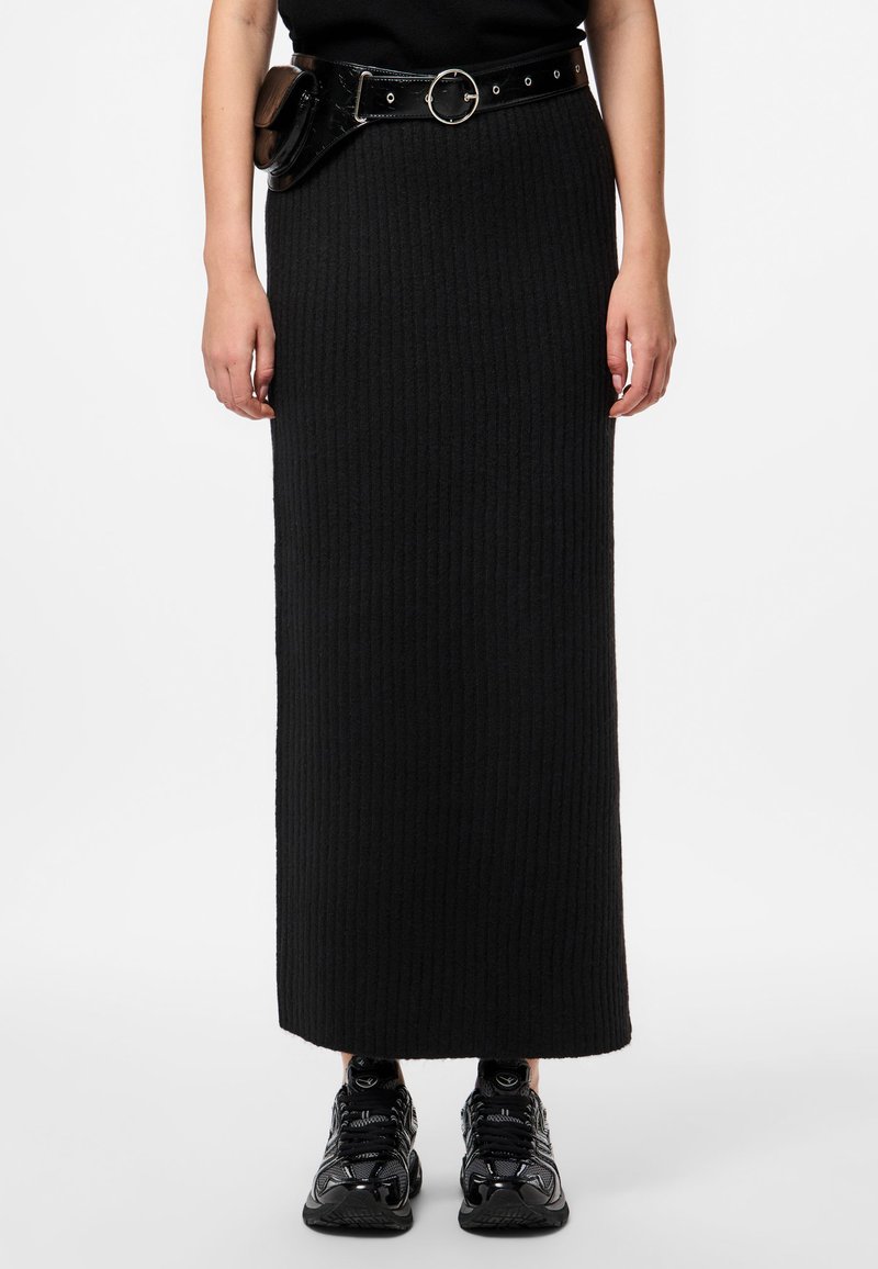 Črna rebrasta maxi krila s prilegajočim silhueto, v kombinaciji z svetlečo črno pasom in črnimi športnimi čevlji. Gladek, teksturiran material.