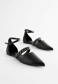 Zapatos planos negros con puntera puntiaguda, material de cuero sintético, tira en el tobillo y detalle de cadena metálica en la parte delantera.