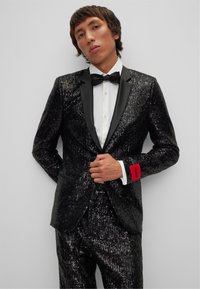 Tuxedo noir à sequins avec revers en satin, associé à une chemise blanche classique et un nœud papillon noir. Étiquette rouge "HUGO" remarquée sur le poignet.