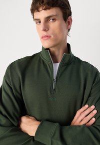 Felpa verde con zip e colletto alto, realizzata in un tessuto testurizzato e con un logo discreto al centro. Dettaglio della zip argentata sul collo.