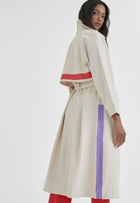 Beżowy bawełniany płaszcz z wcięciem w talii, z czerwonymi i fioletowymi akcentującymi paskami wzdłuż rękawów i boku, oraz wysokim kołnierzem.