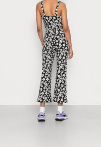 Persona con un top corto de estampado floral en blanco y negro y pantalones acampanados a juego, calcetines blancos y zapatillas moradas y negras, de pie con la espalda visible.