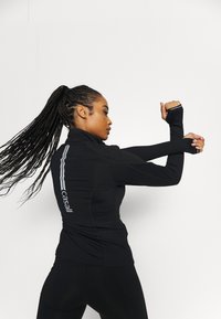 Musta urheilutakki, jossa on pitkät hihat, korkea kaulus ja heijastavat yksityiskohdat selässä. Kangas näyttää sileältä ja joustavalta.