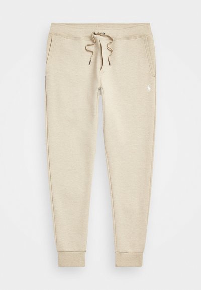 Calças de jogging em bege com formato afunilado, cintura com cordão, bolsos laterais, tornozelos com punho e pequeno logo branco na parte superior da coxa esquerda.