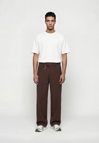 Άνδρας που φοράει απλό λευκό κοντομάνικο μπλουζάκι, χαλαρό καφέ παντελόνι με λουράκι στη ζώνη και λευκά αθλητικά παπούτσια, στέκεται μπροστά σε λευκό φόντο.